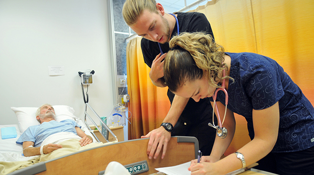 While Dr. Alan Quinn acts as a patient in the background, nursing students Johan Bergenhenegouwen and Ashley Ketler make notes in his chart.