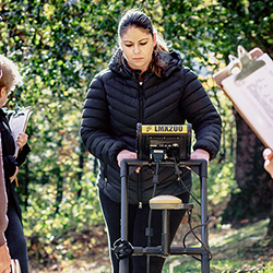 Photo of Dr. Sarah Beaulieu pushing a Ground Penetrating Radar machine in a wooded area, while people with clipboards observe and take notes.