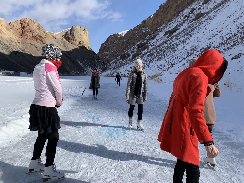 Photo of Chelsea Novakowski and Afghan women skating outdoors on a snow-covered frozen lake, with brown stone cliffs in the background.