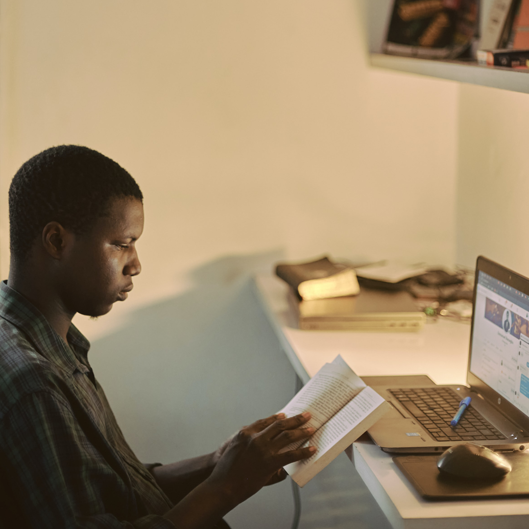 Photo of a young person sitting at their open laptop, studying a book with a pen resting on their keyboard.