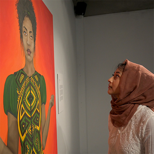 Photo of Faria Firoz looking at her painting hanging in a gallery. The painting shows a Black woman in a green, traditionally patterned dress standing strong with her hands in defiant fists, with a glowing red/orange background and fire in her eyes