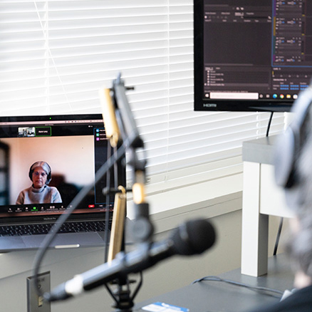 Photo of a studio with professional recording equipment. The focus is on a laptop showing Dr. Jacqueline Nolte on a Zoom call, while in the foreground, Dr. Martha Dow speaks into a microphone.