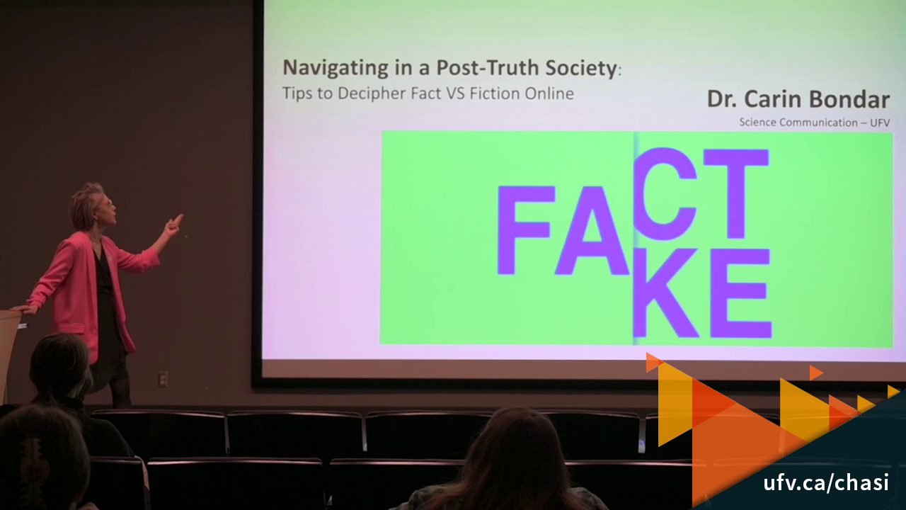 Photo of Dr. Carin Bondar presenting on stage. The screen shows a slide which reads "Navigating in a post-truth society: tips to decipher fact vs fiction online. Dr. Carin Bondar, Science Communication, UFV." A large graphic shows the letters FA, with the second half of the word as both CT (spelling fact) and KE (spelling fake).