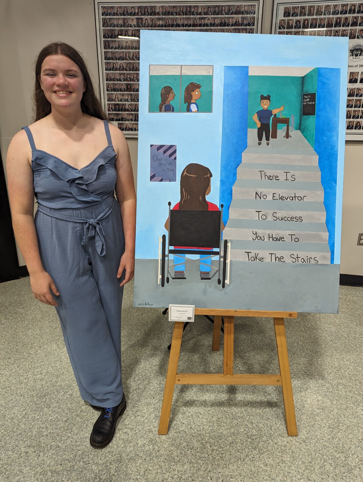 Photo of Lexis De Meyer standing next to a painting. The painting shows a person in a wheelchair at the bottom of a staircase leading to a classroom. The stairs have writing on each step, reading "there is no elevator to success, you have to take the stairs."