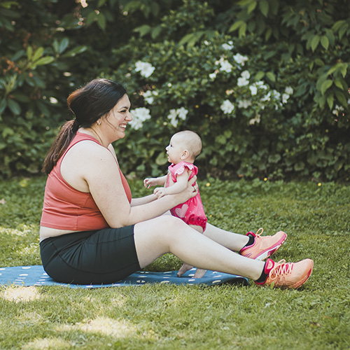 Thumbnail photo of a mother sitting on an exercise mat outdoors, holding her baby upright and smiling.