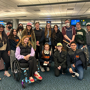 Group photo of 15 members of the CHASI team posing in an airport at the end of the trip home. Many are wearing navy blue sweatshirts with CHASI written on them in orange letters.
