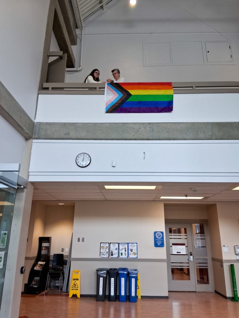 Photo of two people hanging a pride flag from the second story of a open grand hall at UFV