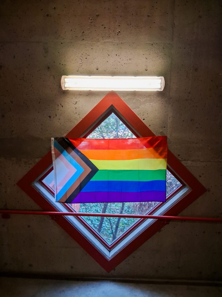 Photo of a pride flag hanging in front of a bright window in a dark staircase