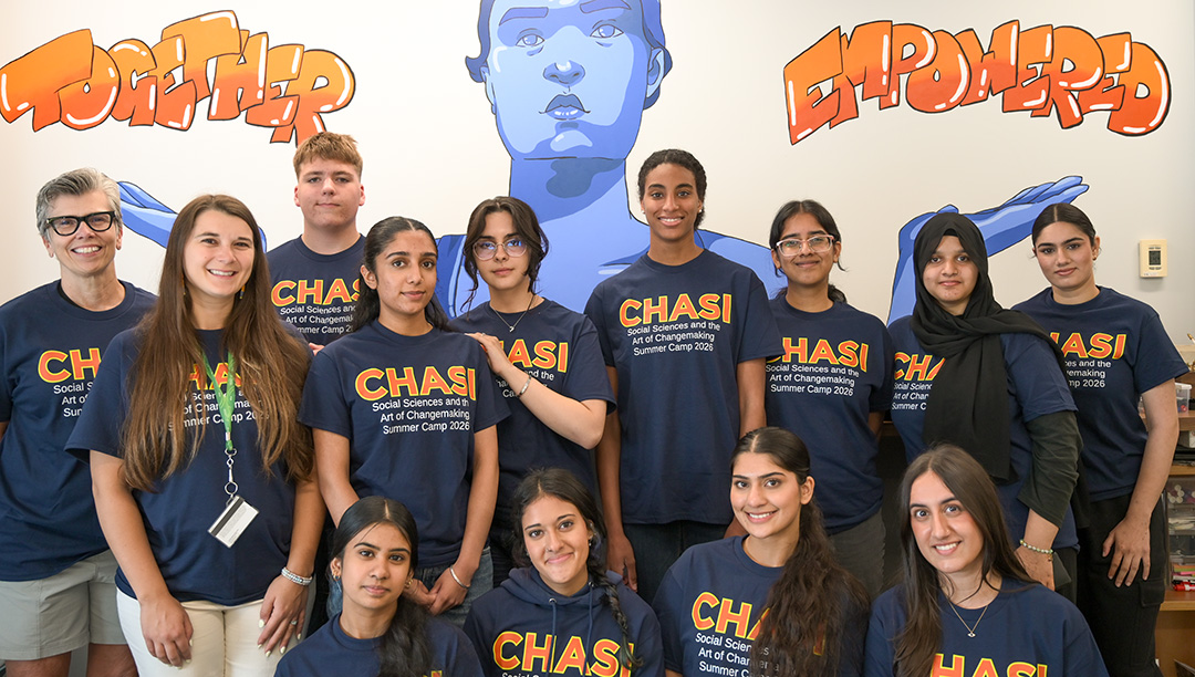 Group photo of the students and leaders of the CHASI summer camp, posing in matching summer camp t-shirts in front of CHASI's "Together Empowered" mural.