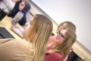 Close-up of two students seated in a UFV classroom, listening to a presentation during a CMNSA meeting, with water bottles and laptops on the desk.