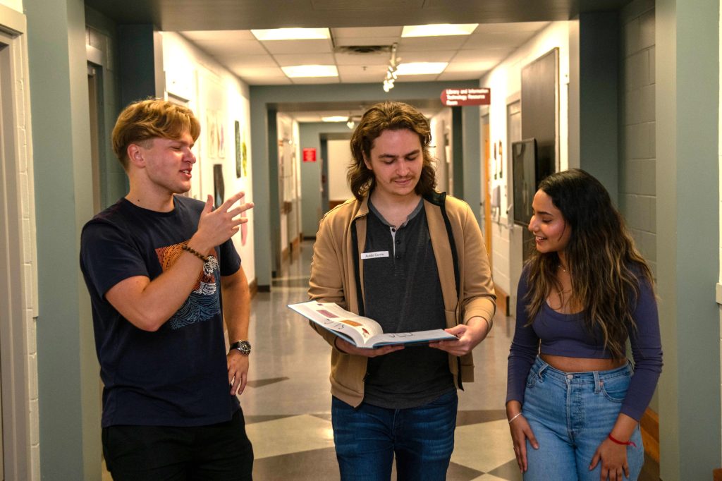 3 students walking in a hallway talking and the one in the middle is looking down at an open textbook.