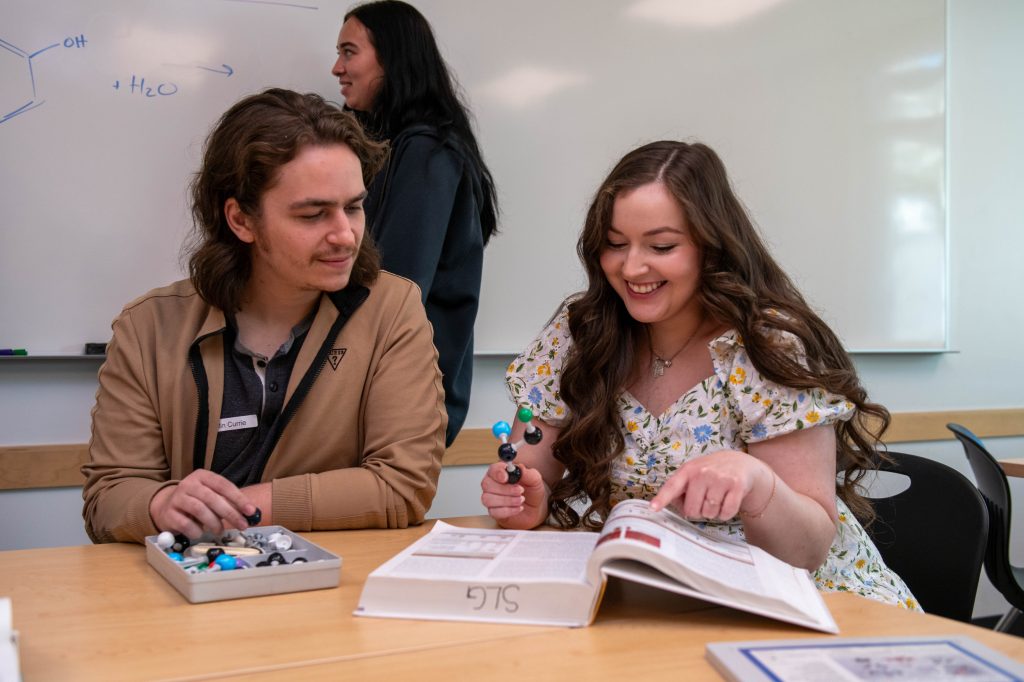 Two students looking at a textbook smiling and holding atom models.