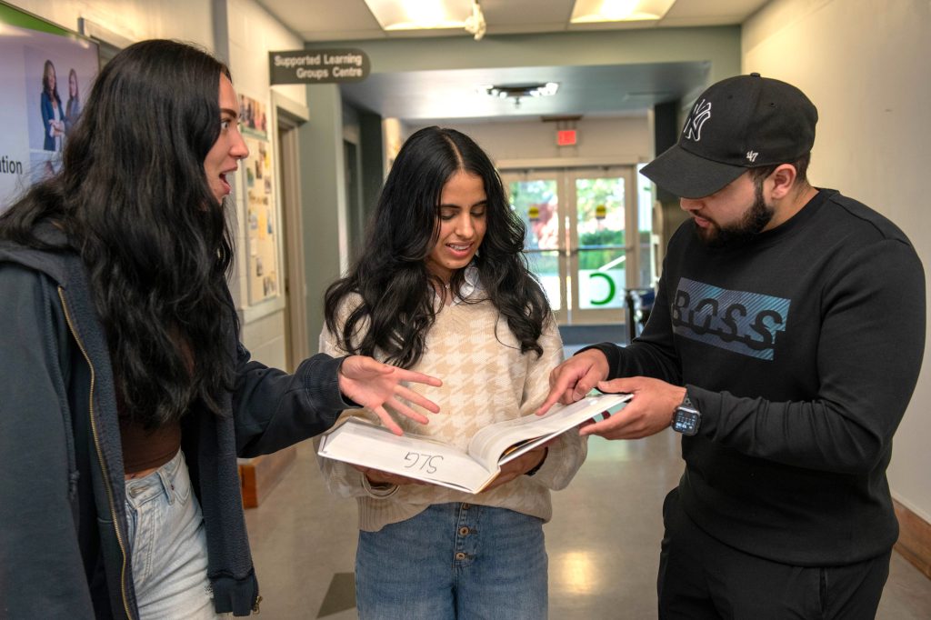 3 students standing in a hallway all looking at the textbook and one student looks shocked.