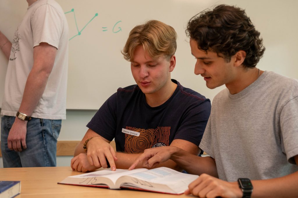 2 students sitting in a classroom pointing to the same spot at an open textbook.