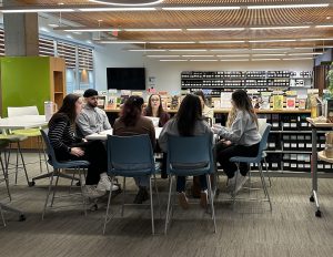 7 students sitting in chairs at a square desk discussing and looking engaged