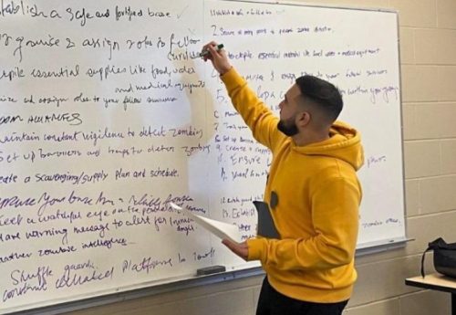 Harman in a yellow hoodie writing on a whiteboard